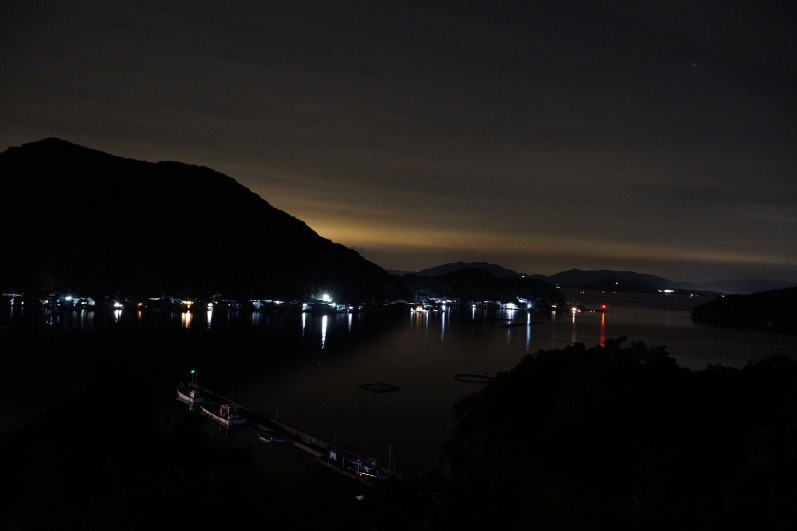 Ine Bay at night from the Michi no Eki hilltop, fishing village lights reflecting on the dark water, mountains silhouetted against the glow