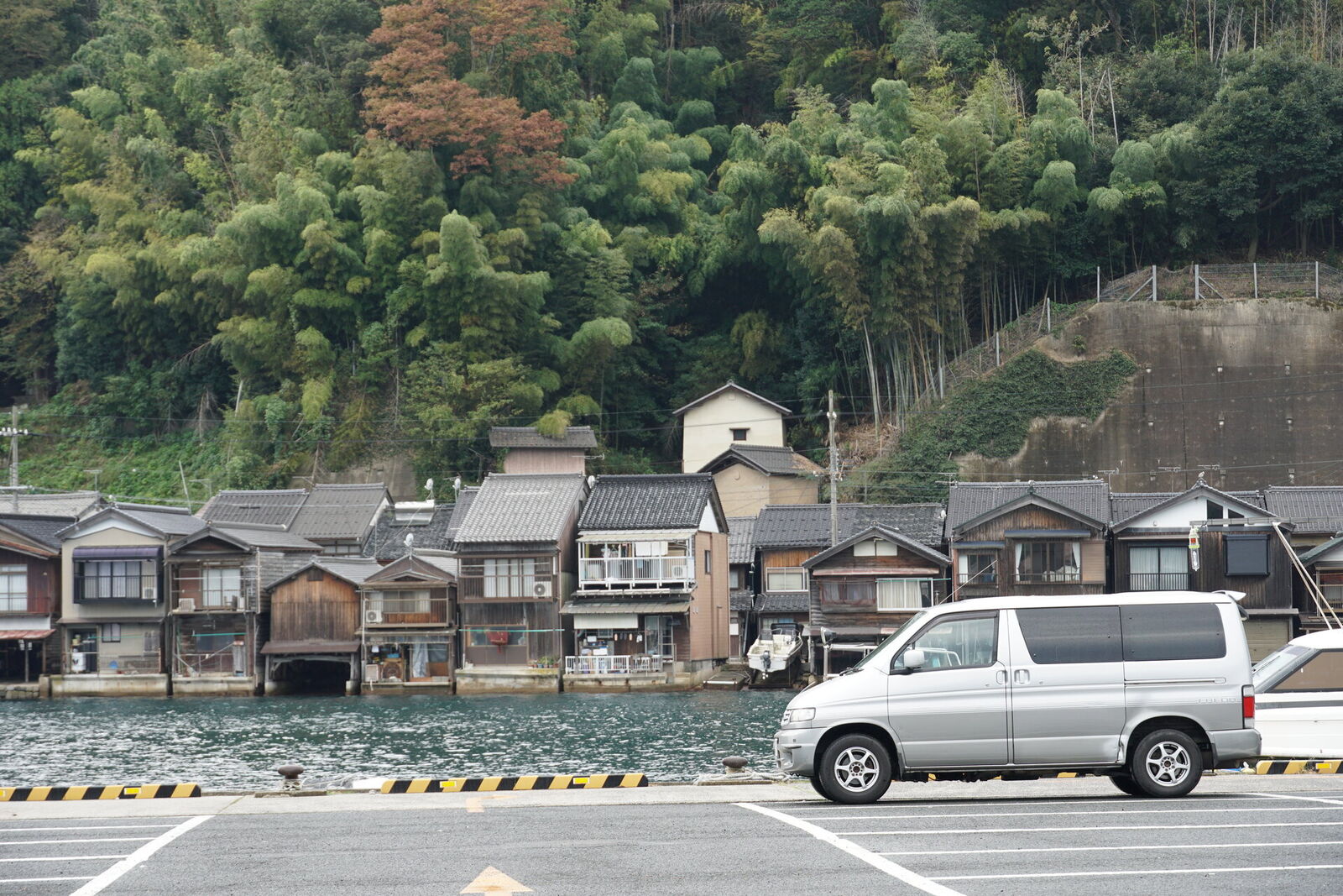 Mazda Bongo campervan parked at Ine harbor with traditional funaya boathouses lining the bay and forested hillside behind