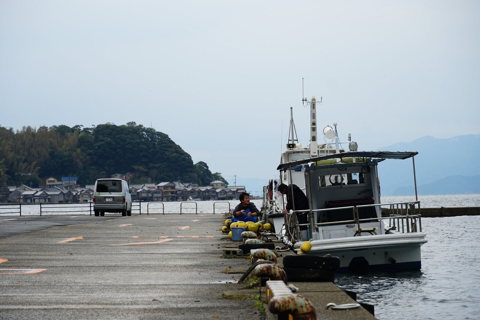 A fisherman sitting by his boat on the pier, the bay and funaya village stretching out behind him in the afternoon light