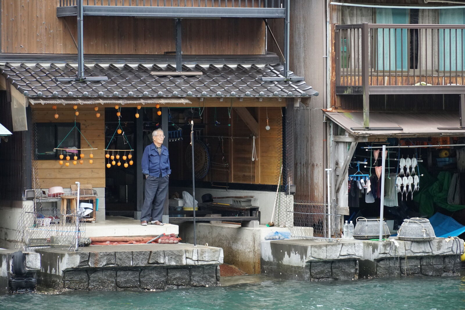 An elderly fisherman standing in the doorway of his funaya with orange fishing floats hanging behind him and nets drying nearby