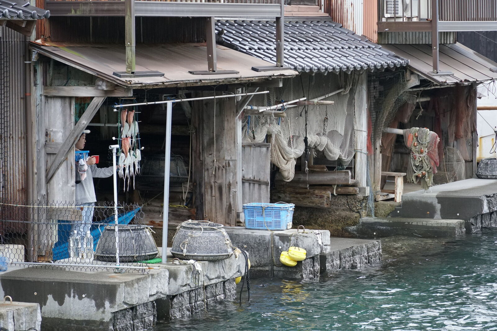 Fisherman hanging fresh catch to dry outside his weathered funaya, with fishing traps and nets stacked at the waterline