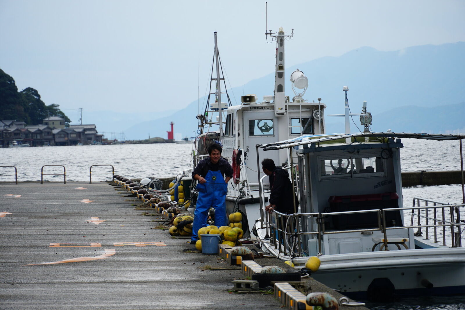 Fisherman in blue overalls working at the pier, his boat moored alongside, misty mountains across the bay