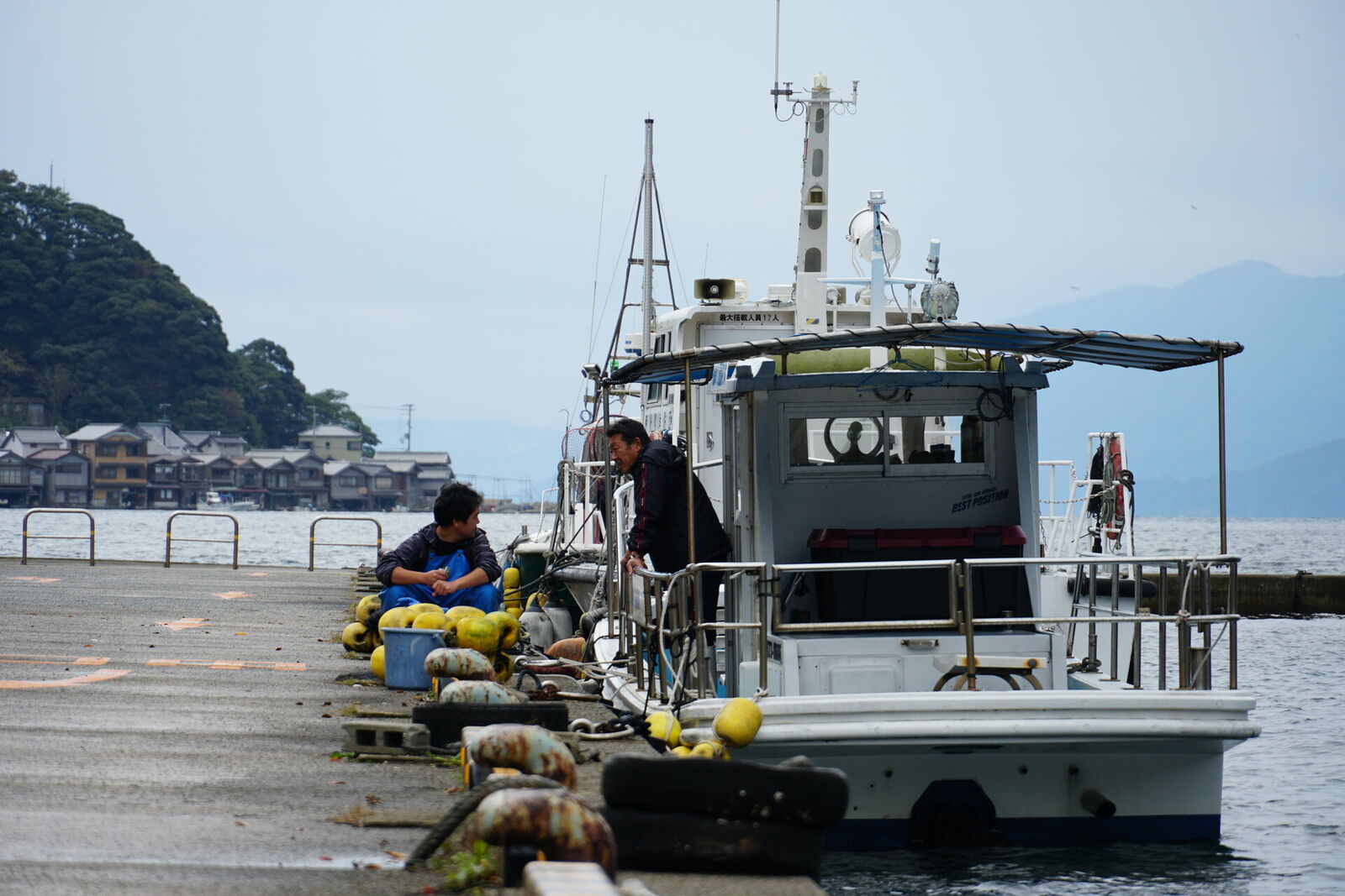 Two fishermen at the concrete pier tending to their fishing boat, with the bay, funaya, and mountains forming the backdrop
