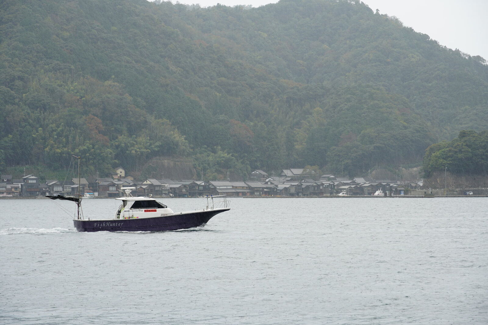 A blue fishing boat crossing the bay with funaya village in the misty background, showing the daily rhythm of Ine's fishing community