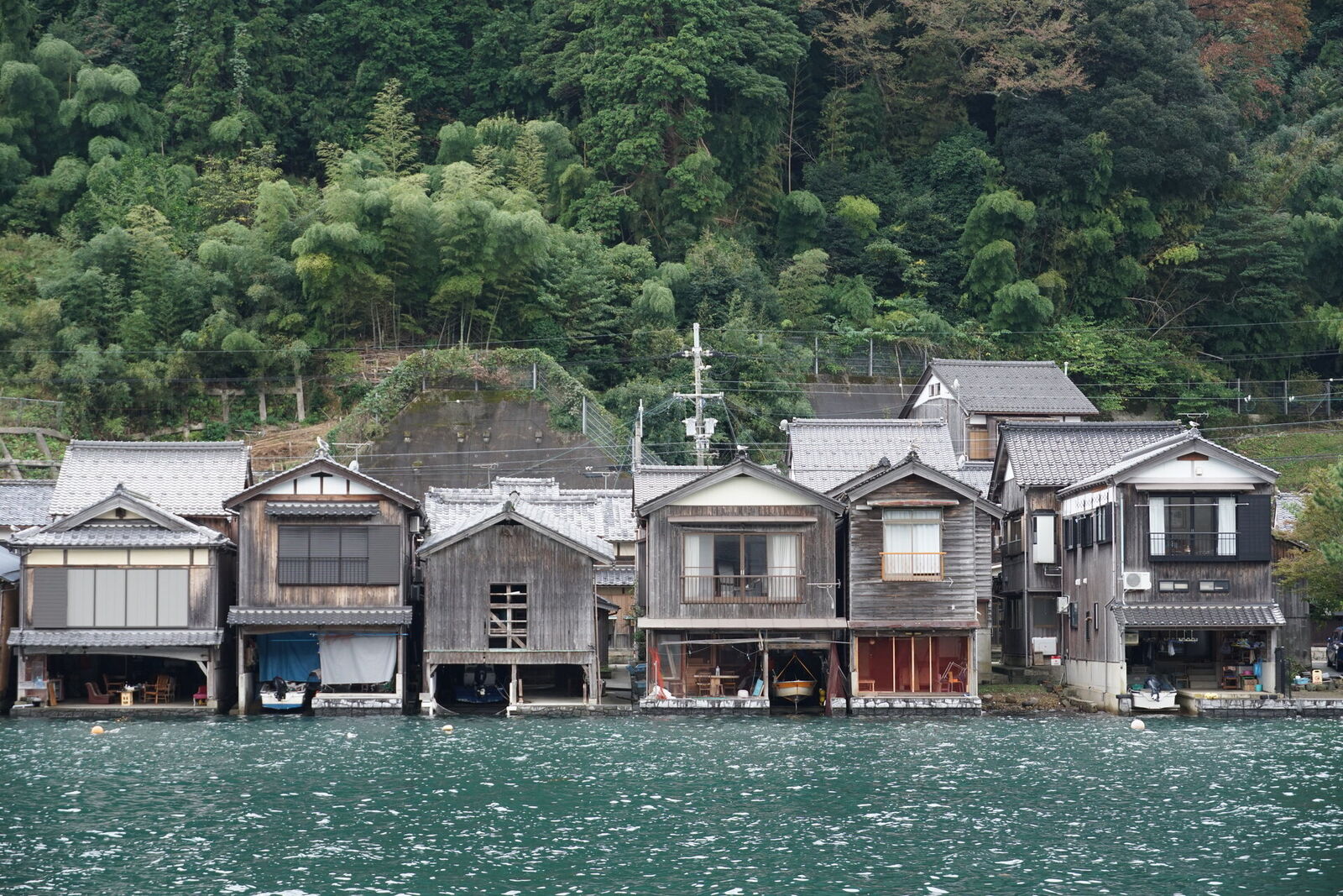 Close-up of funaya boathouses from the water, showing the distinctive two-story architecture with boat garages below and living quarters above, bamboo grove behind