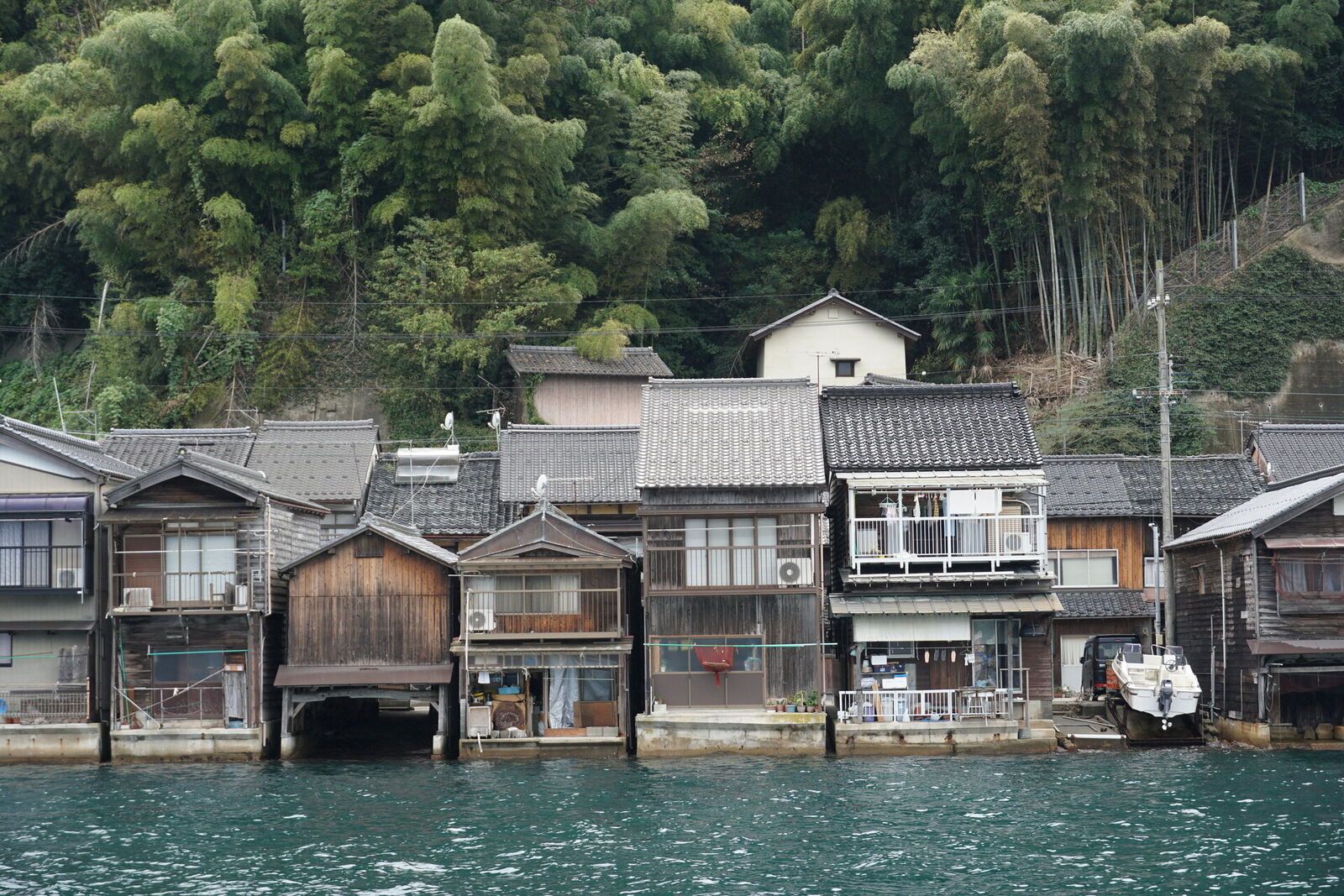 Classic funaya row seen from the water, wooden boathouses with their characteristic boat garages opening to the bay, bamboo forest on the hillside behind