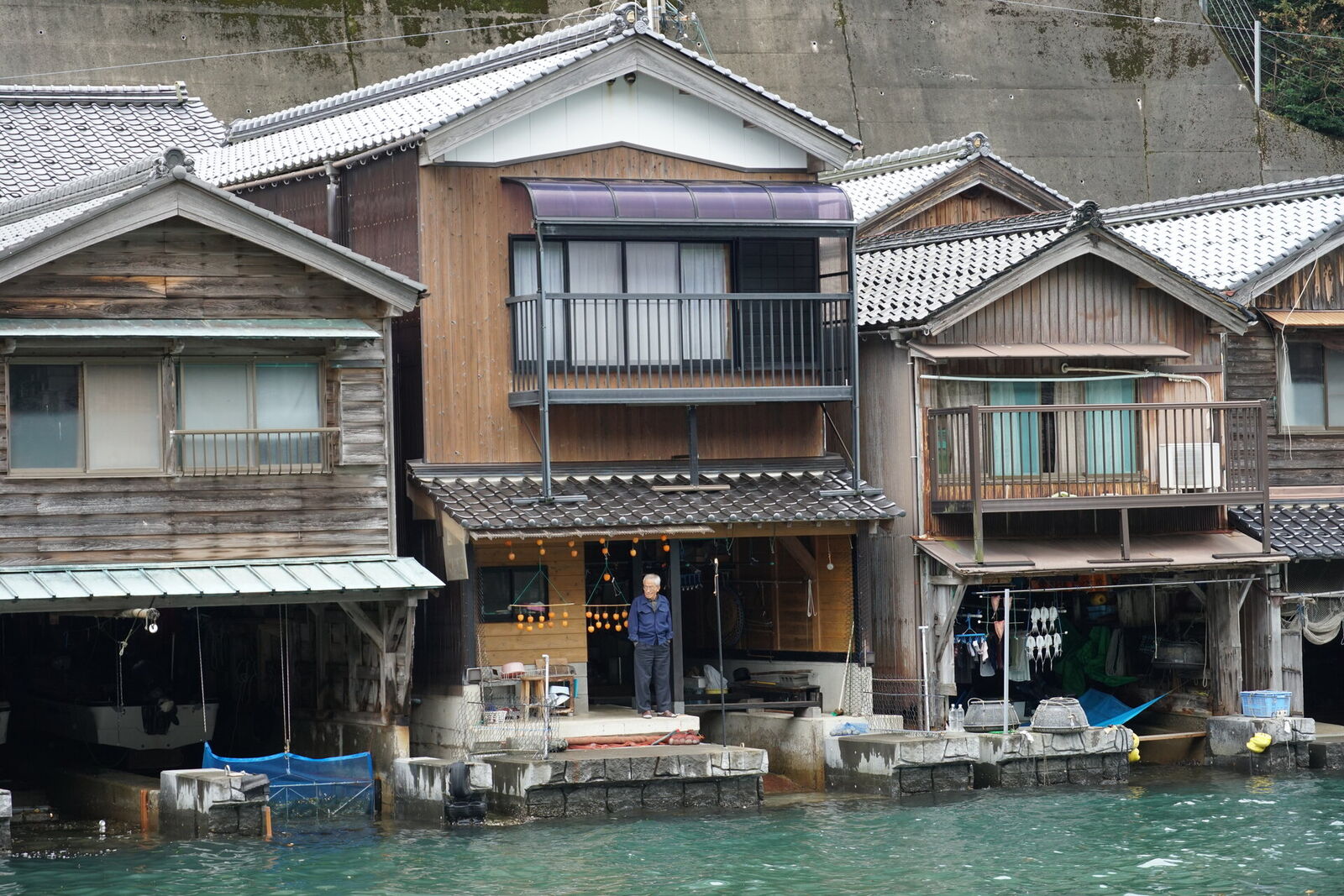 A man standing in front of his funaya with string lights, looking out across the water, boats at the waterline below