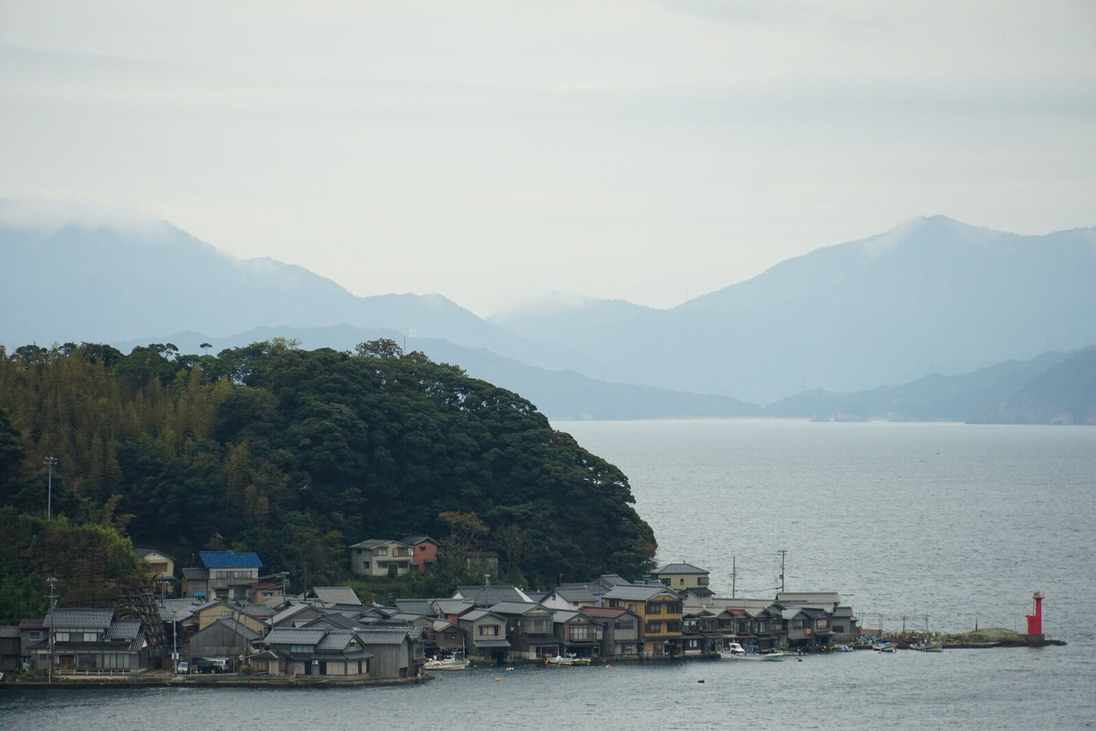 Ine village panorama with funaya lining the coast, a red lighthouse at the bay entrance, and layered mountain ridges fading into the mist