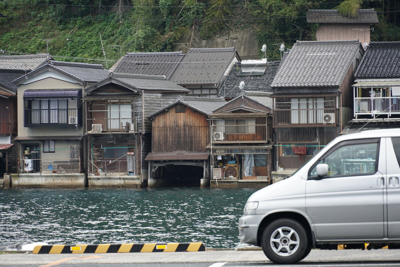 Campervan driving past traditional funaya boathouses on the water at Ine fishing village