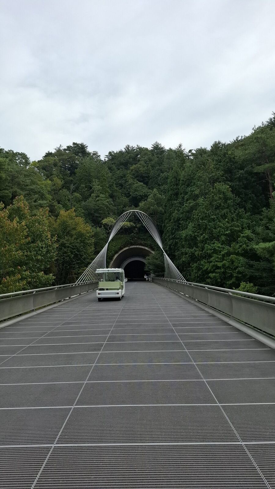 The full suspension bridge stretching from the tunnel mouth toward the museum, cables sweeping upward, an electric cart crossing, forested mountains on all sides