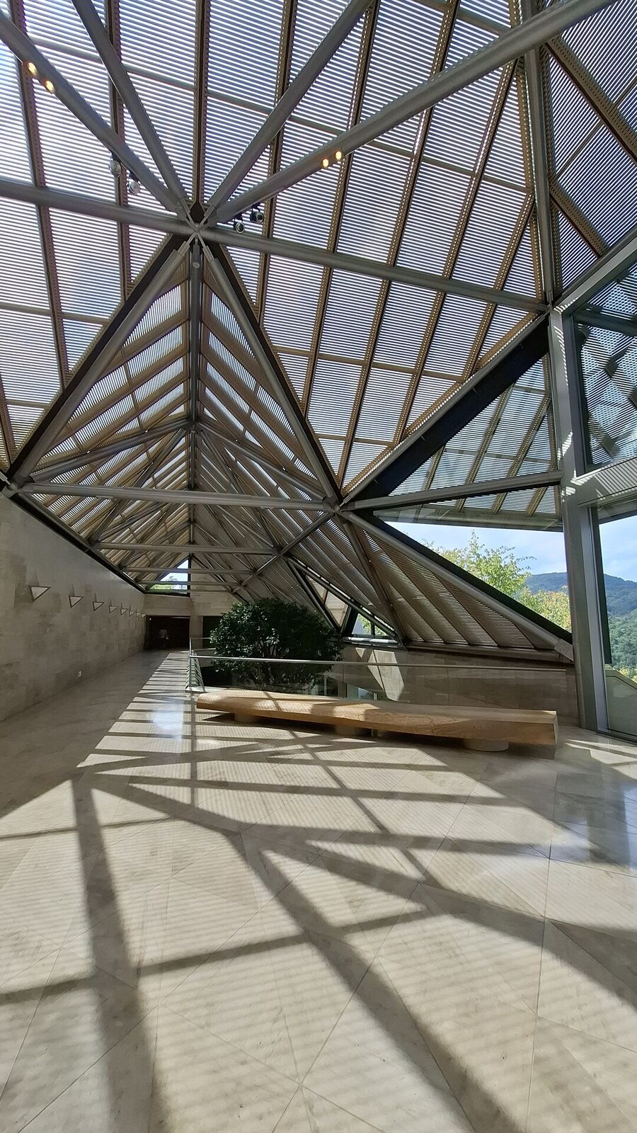 The glass roof casting dramatic geometric shadows across the limestone corridor, steel beams and wooden louvers creating a play of light and pattern