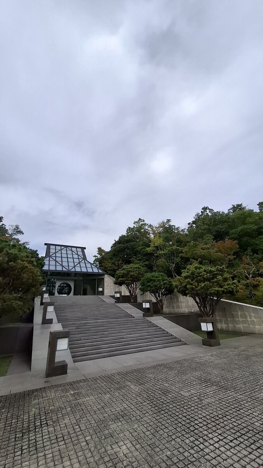 The museum facade from below, stone steps flanked by bollard lanterns leading up to the glass and steel roof structure emerging from the hillside