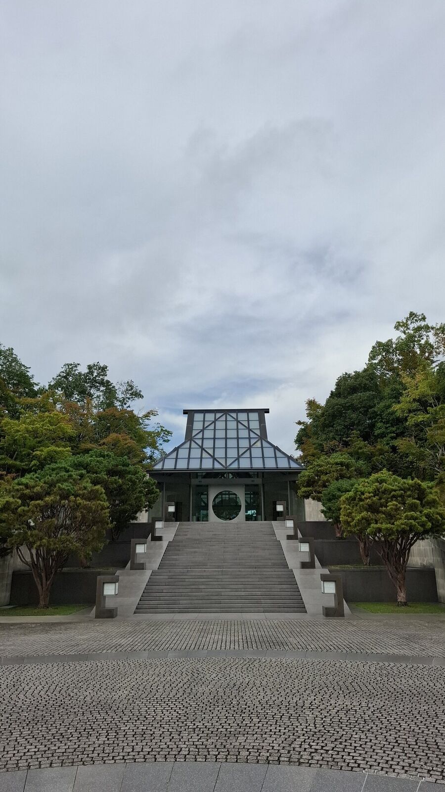 The iconic frontal view of Miho Museum, symmetrical grand staircase ascending to the glass entrance with its circular moon window, framed by sculpted pines