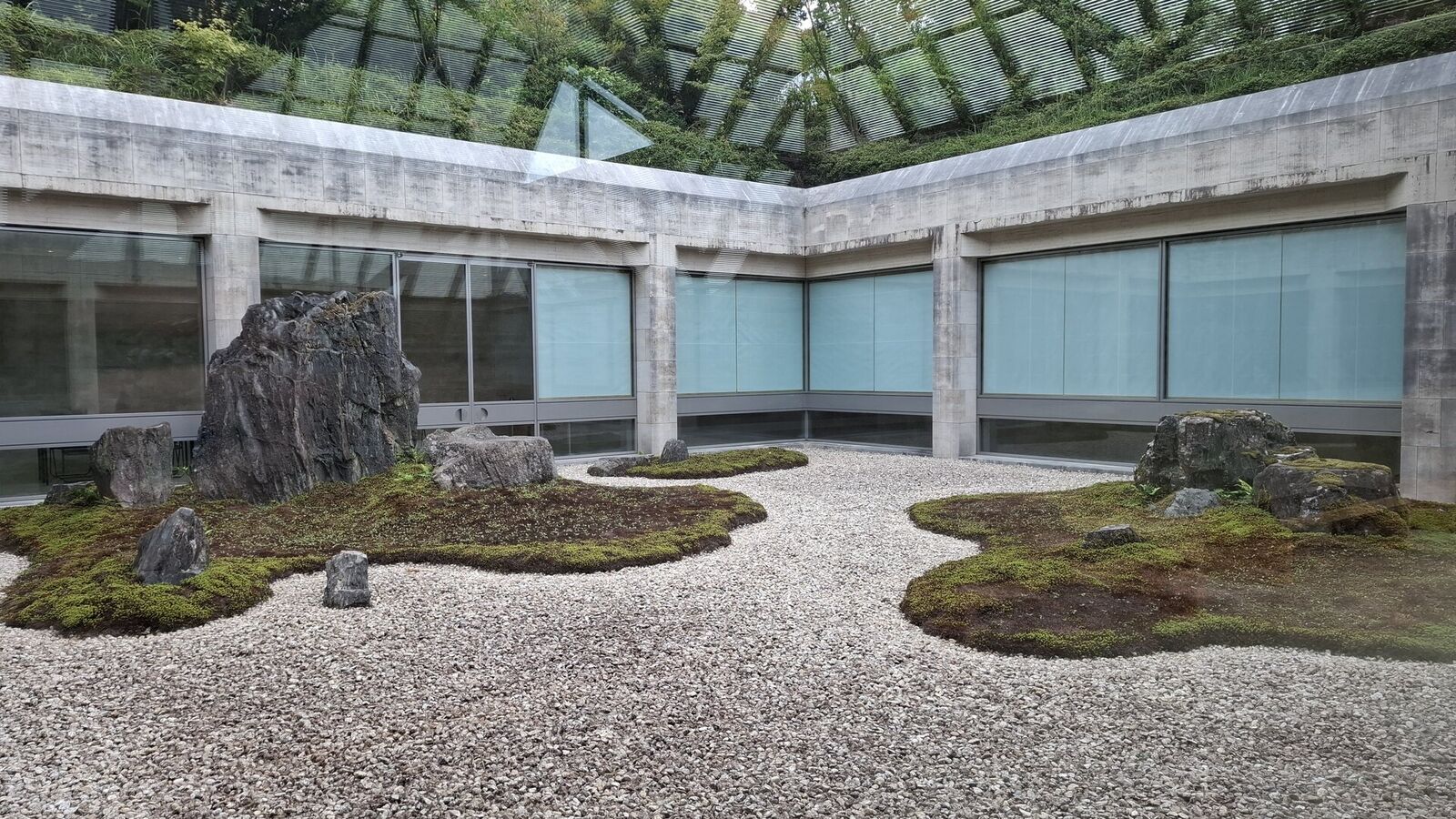 The sunken zen garden courtyard with carefully placed rocks, raked gravel, and moss islands, enclosed by concrete walls with blue-green frosted windows