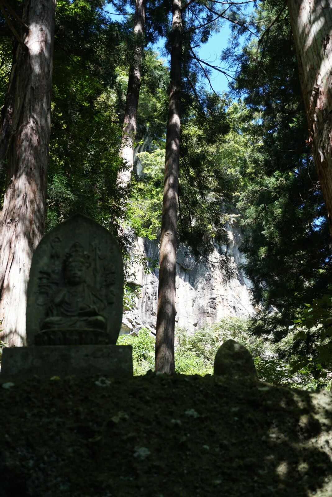 Stone Buddha figure carved into sandstone, moss-covered and weathered, standing among the forest graves