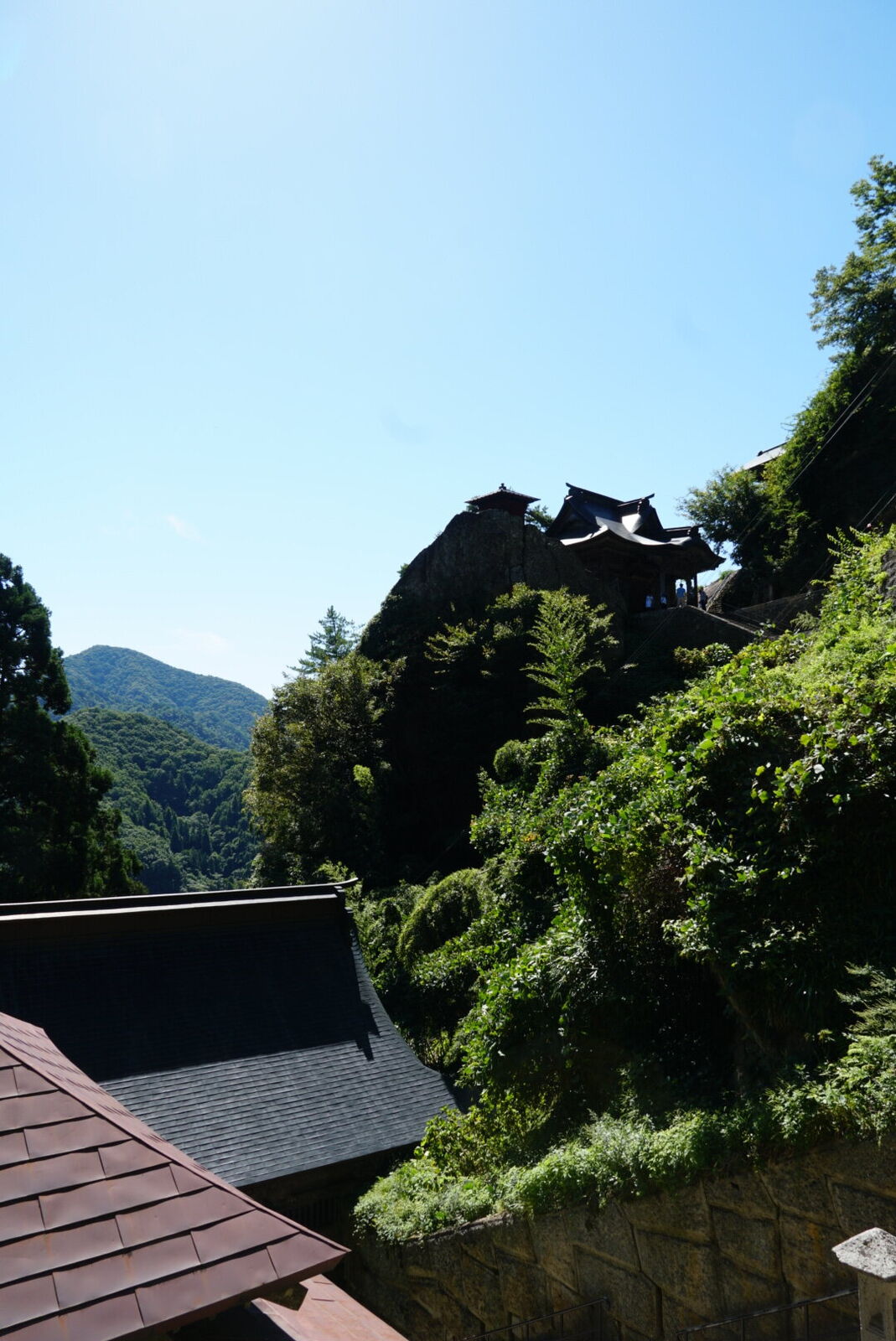 Multiple temple buildings clinging to the cliff face, wooden structures on narrow ledges with green mountains and blue sky behind