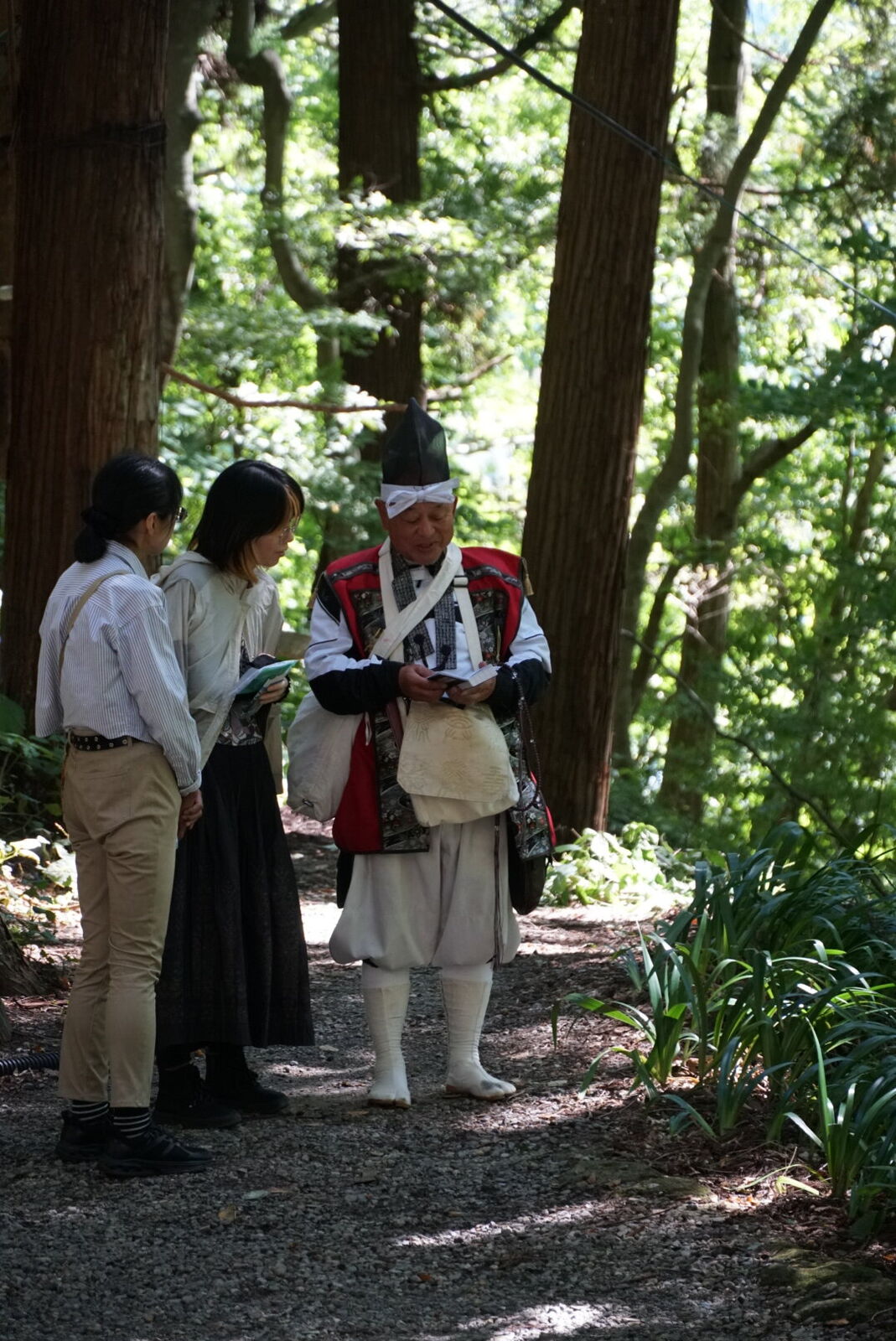 A Yamabushi mountain ascetic in traditional white robes and black hat speaking with a Japanese couple on the forest path