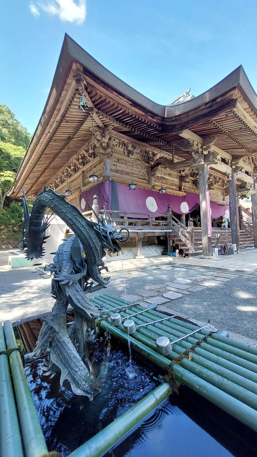 The dragon temizuya at Nariaiji with the main hall rising behind it, purple curtains and intricate wooden joinery visible under a summer sky