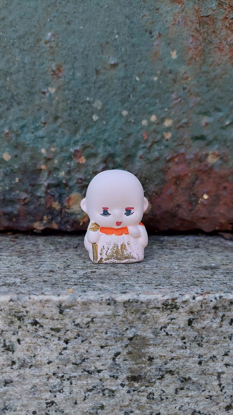 A tiny ceramic Jizo figure sitting on the edge of a stone basin at Nariaiji, white porcelain with gold detailing and a serene smile