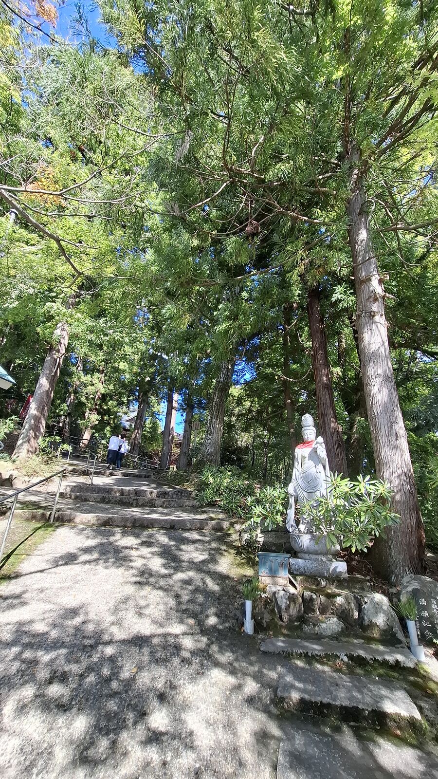 A stone Jizo statue with red bib standing at the base of stone steps in a cedar forest, the path leading up to Nariaiji temple