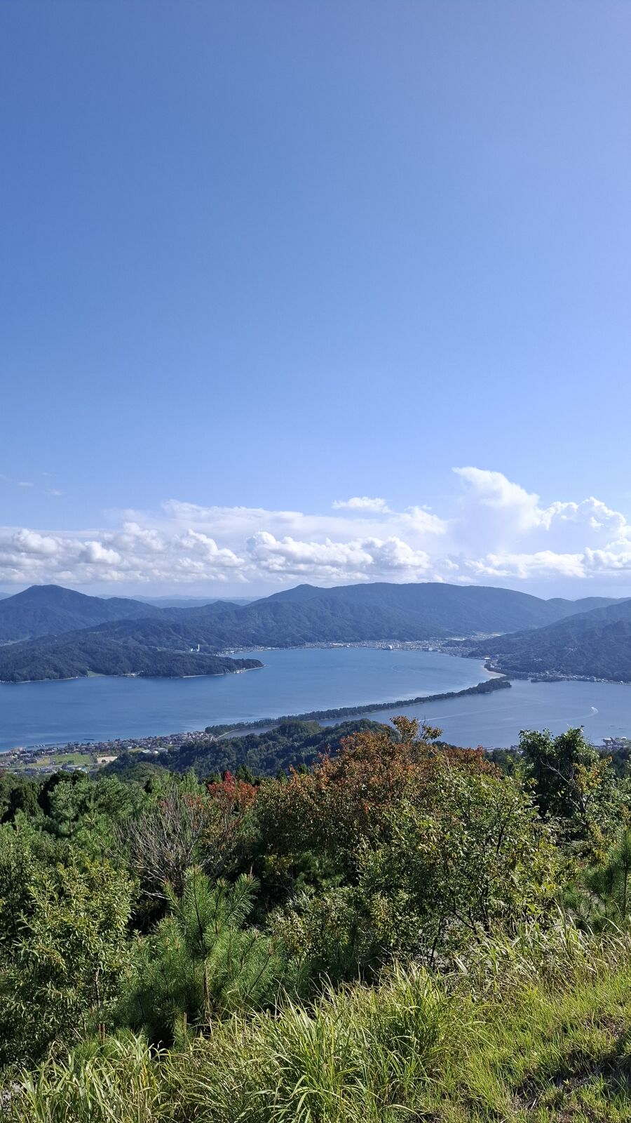 View from Nariaiji looking across the bay toward distant mountains, autumn-tinged vegetation in the foreground, clouds above the peaks
