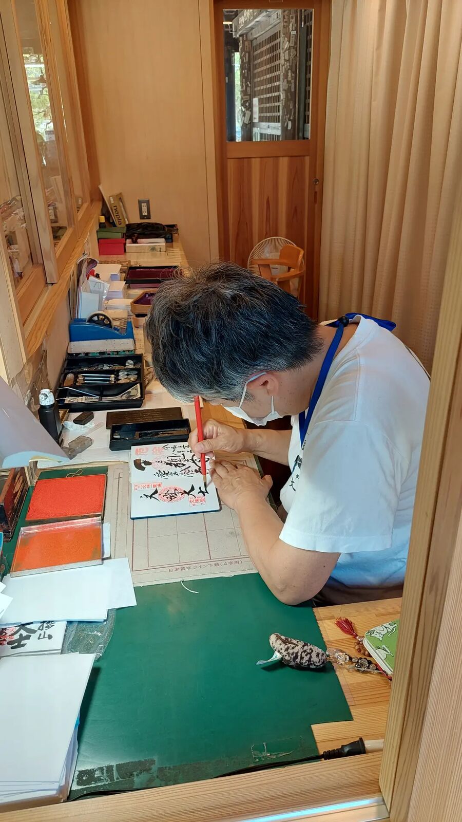 A temple calligrapher writing Goshuin in a visitor's stamp book, bent over his work at a wooden desk surrounded by brushes and ink