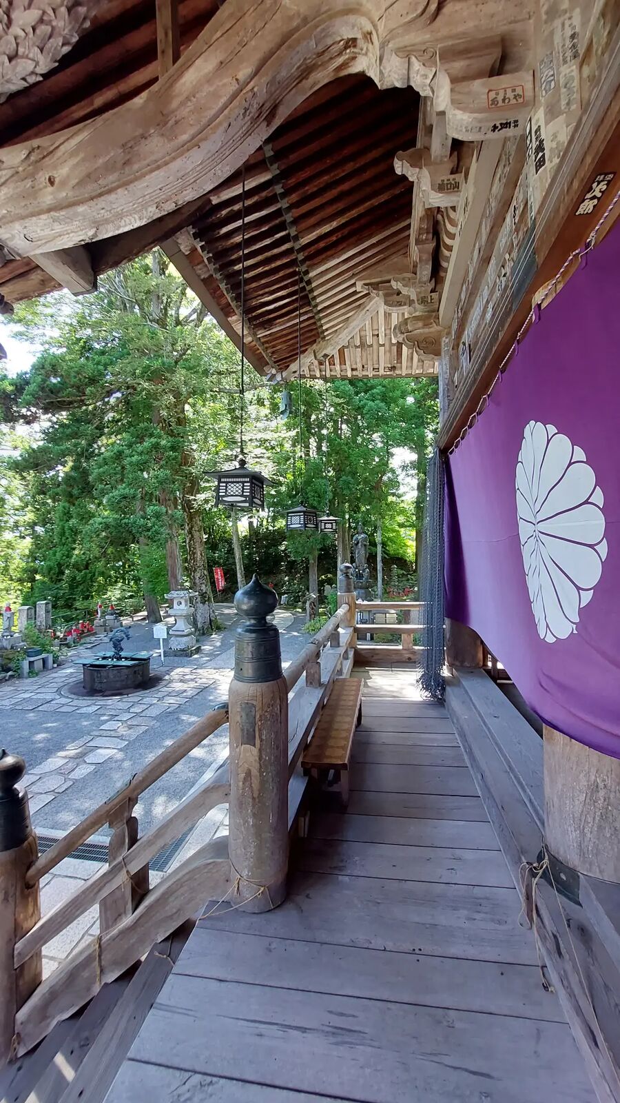 The wooden veranda of Nariaiji temple stretching into the distance, purple ceremonial curtain with chrysanthemum crest hanging beside ornate roof brackets, a bronze lantern in the shade