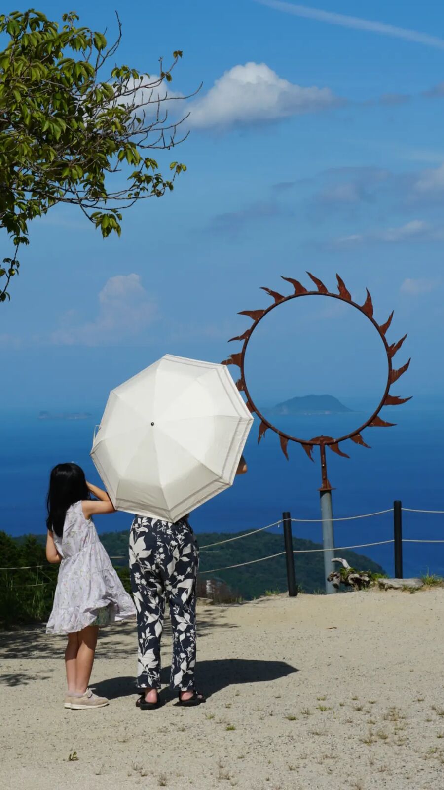 A woman and child with a white parasol standing at the sun sculpture viewpoint above Nariaiji, the Sea of Japan and distant islands visible through summer haze