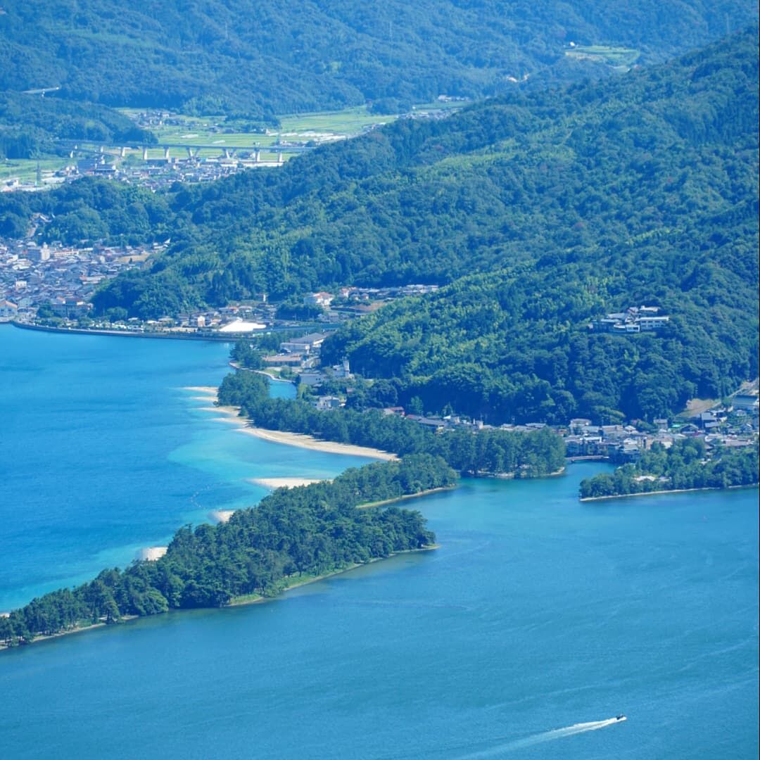Aerial view of the Amanohashidate sandbar from the mountaintop above Nariaiji, the pine-covered strip curving across turquoise water with a boat leaving a white wake