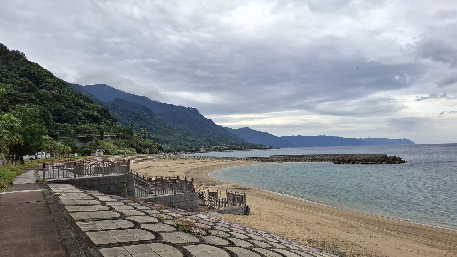 Coastal panorama on the way to Cape Sata, a stone-paved path along a sandy beach with forested mountains rising steeply from the water under heavy clouds