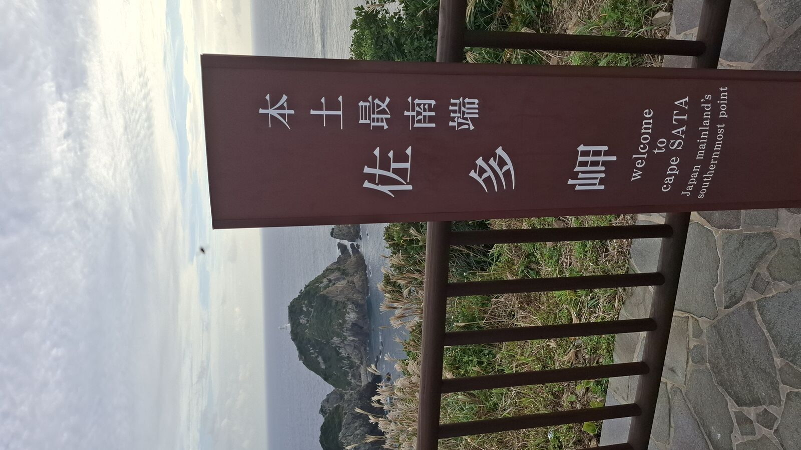 Wooden sign reading Welcome to Cape SATA with dramatic cliffs and ocean visible behind, subtropical plants framing the scene