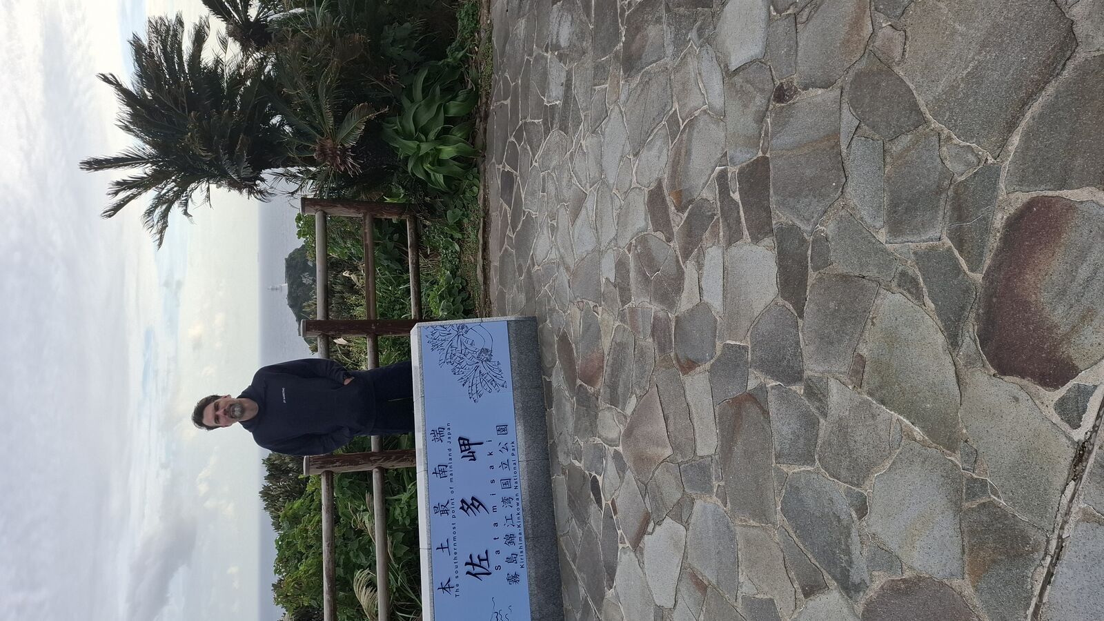 Kevin standing next to the 佐多岬 stone marker sign with a palm tree behind, the official southernmost point monument