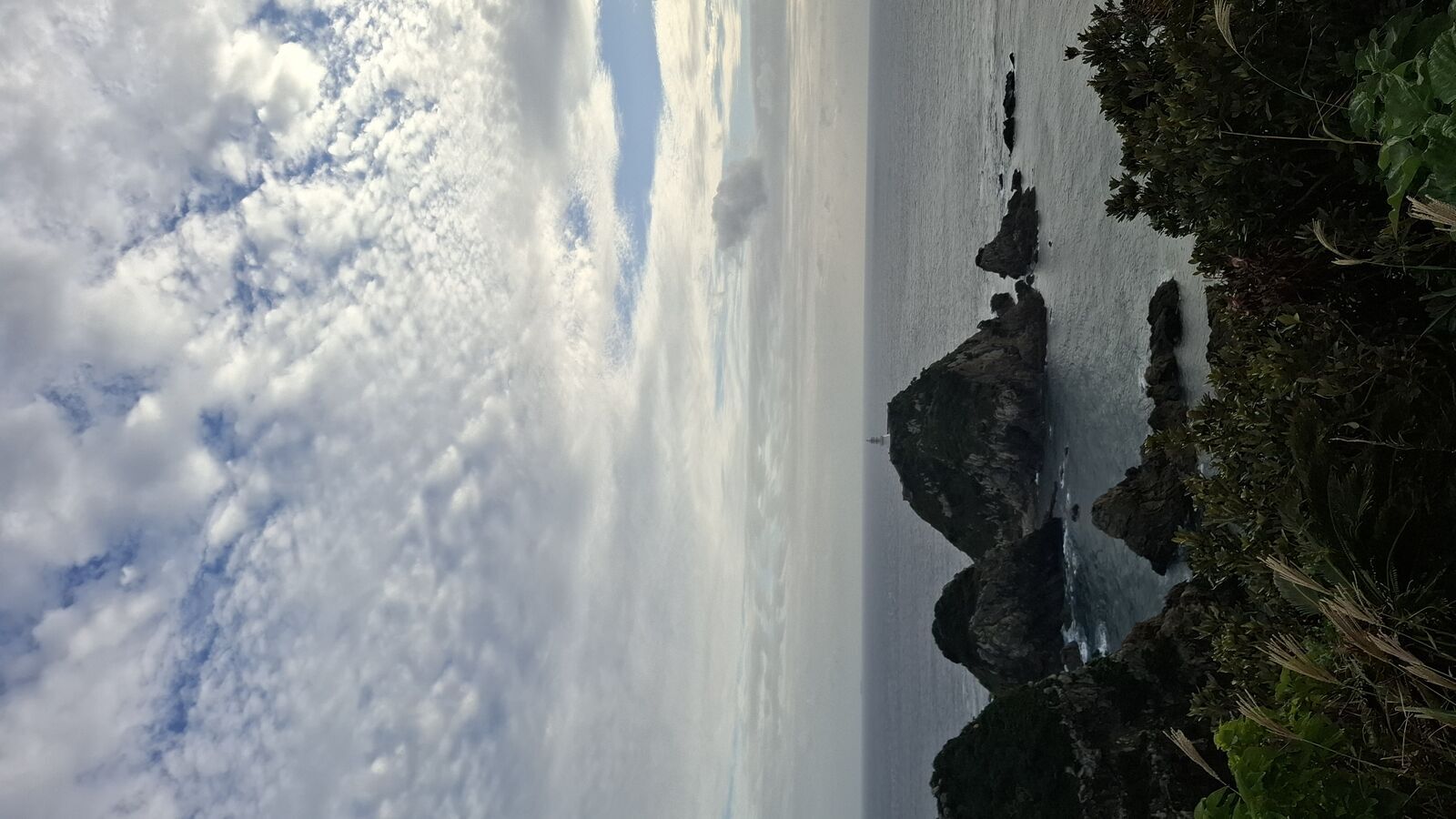 Dramatic cliff panorama with the lighthouse rock in the distance, deep blue ocean, waves breaking white against the rocks below