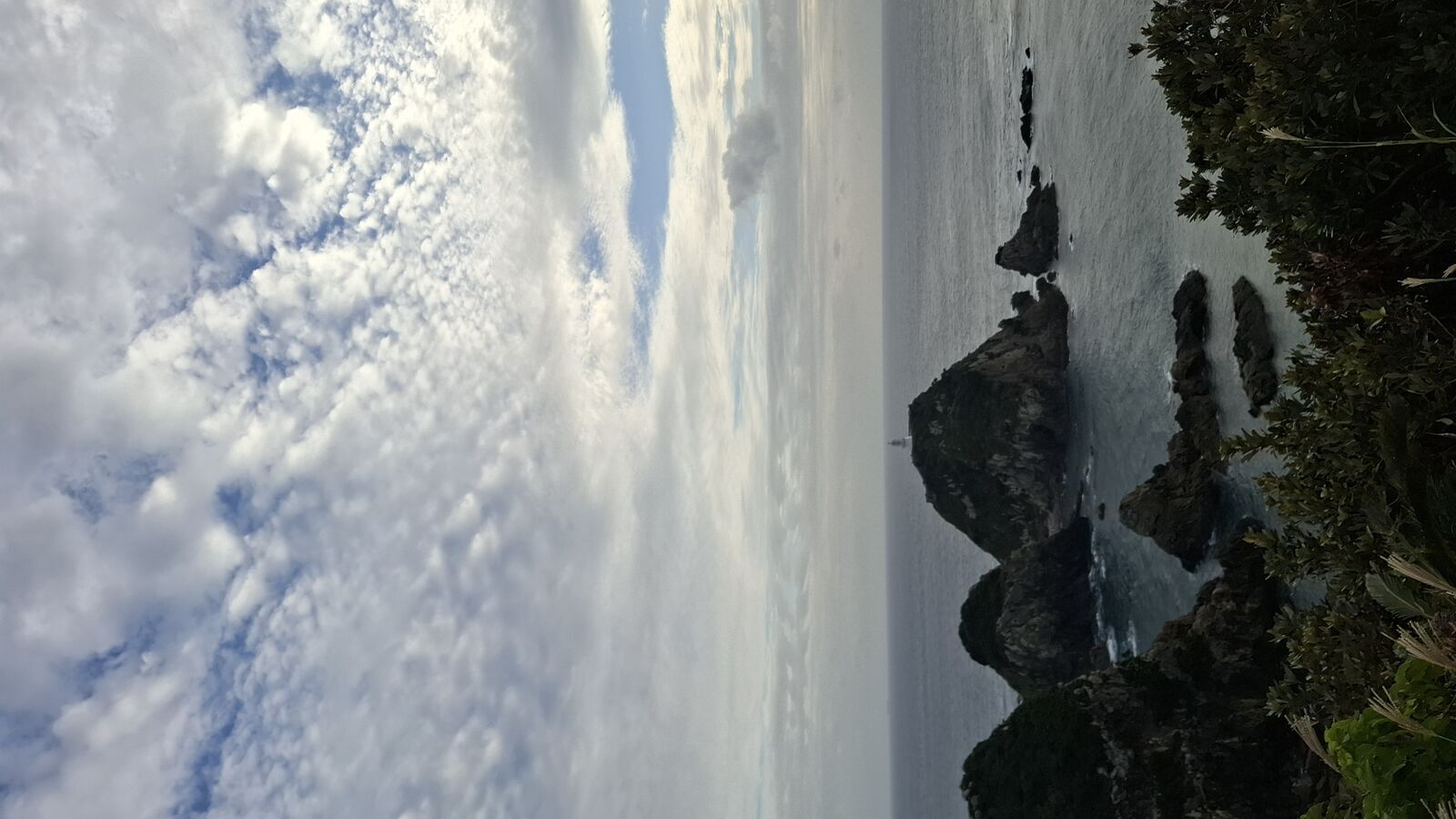 Wide view of the cape's rugged coastline, rocky outcrops in the ocean, the lighthouse visible on its island far below
