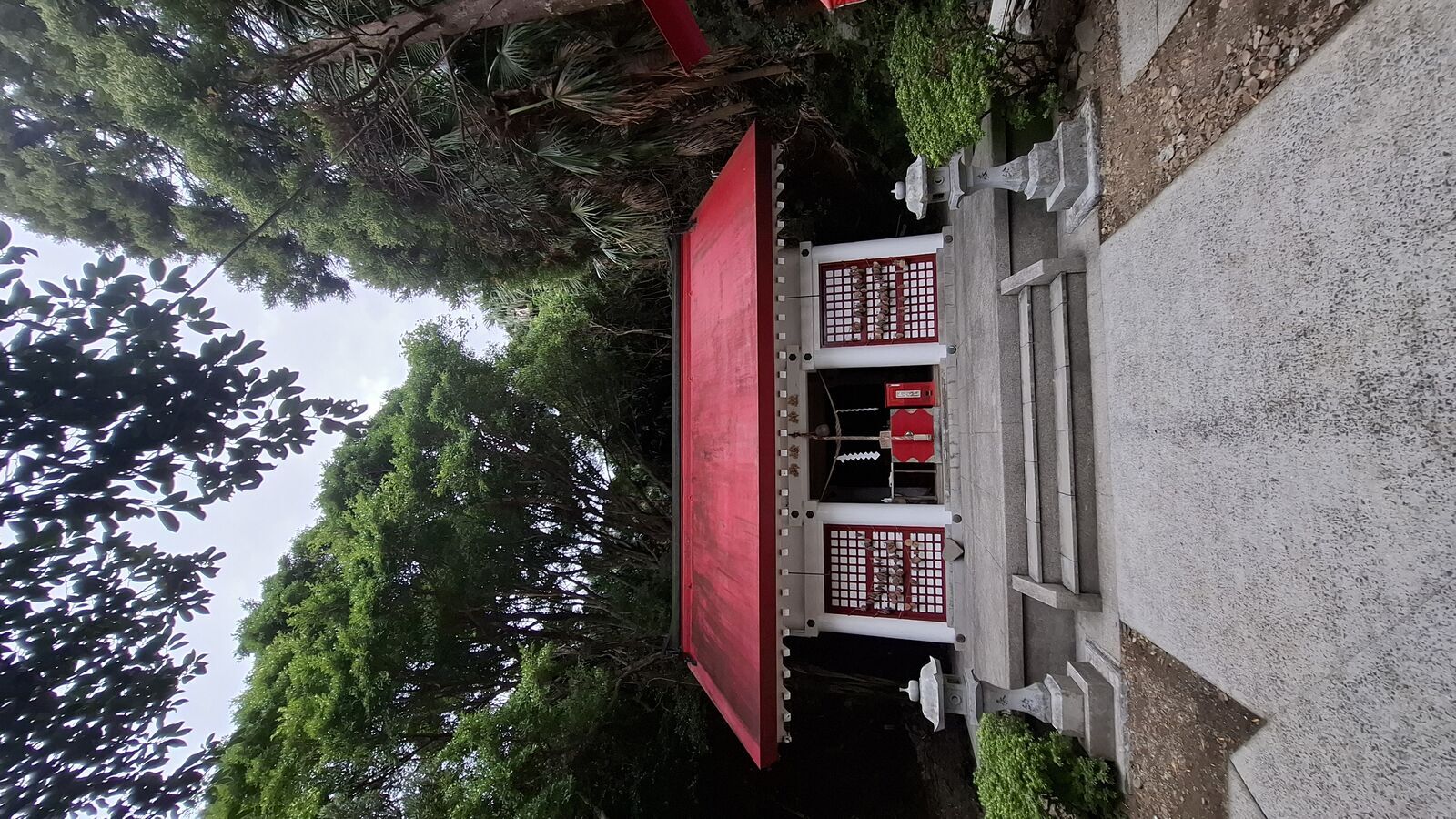 Small red shrine building with white and red lattice details, surrounded by subtropical trees and palm fronds