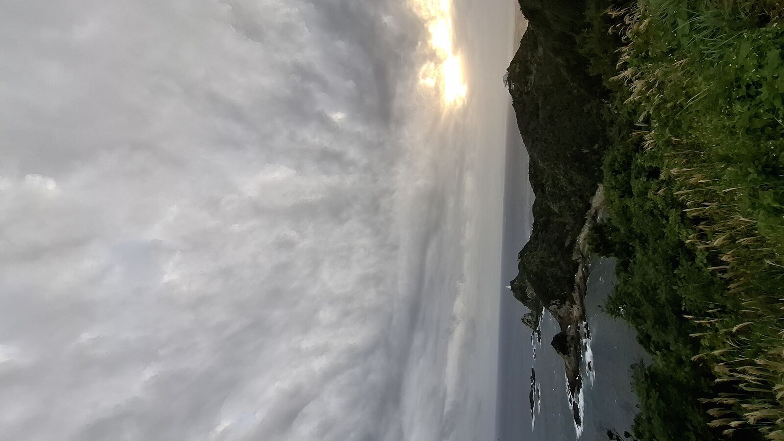 Golden sunlight breaking through clouds onto the cliff face, warm light illuminating the rocks and subtropical vegetation