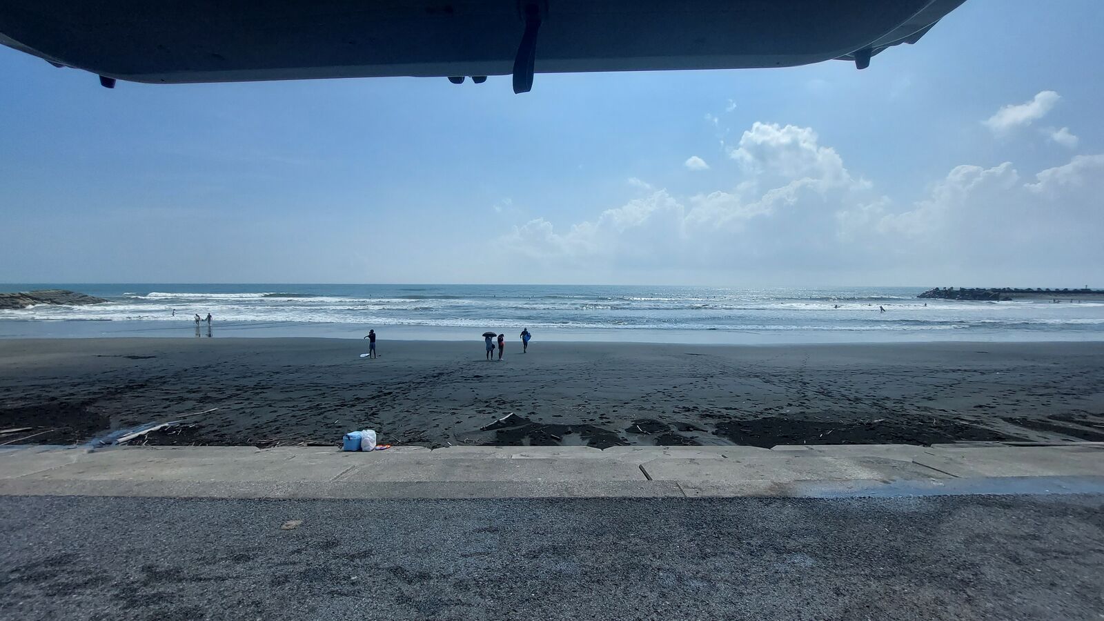 View from underneath the van's open trunk: beach, blue sky, swimmers in the distance, a blue cooler on the sand