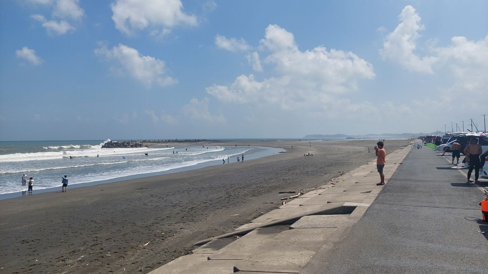 Long view down Kujukuri Beach: concrete seawall, dark sand, swimmers, tetrapod breakwaters, and the coastline disappearing into summer haze