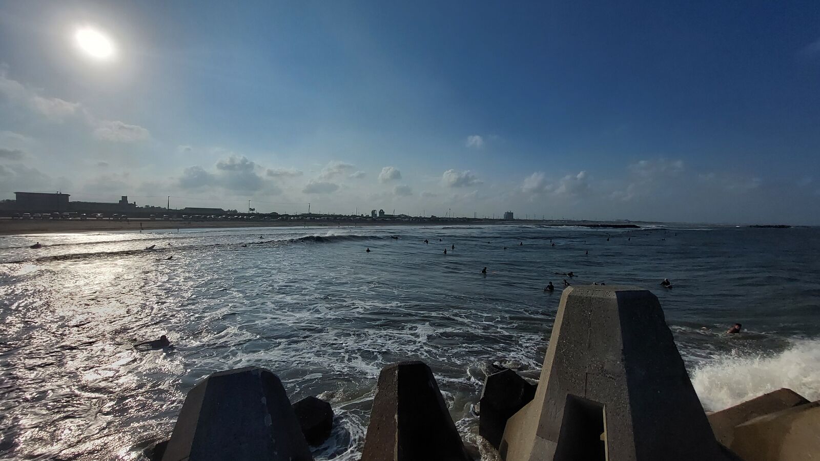 Afternoon sun over the Pacific seen through concrete tetrapod breakwaters, swimmers scattered in the sparkling water, city skyline in the distance