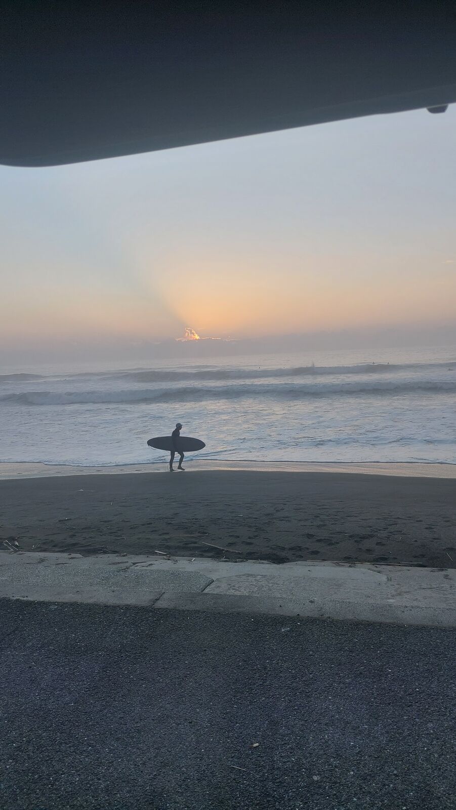 Lone surfer with board silhouetted against dawn sky, seen from inside the van through the open back