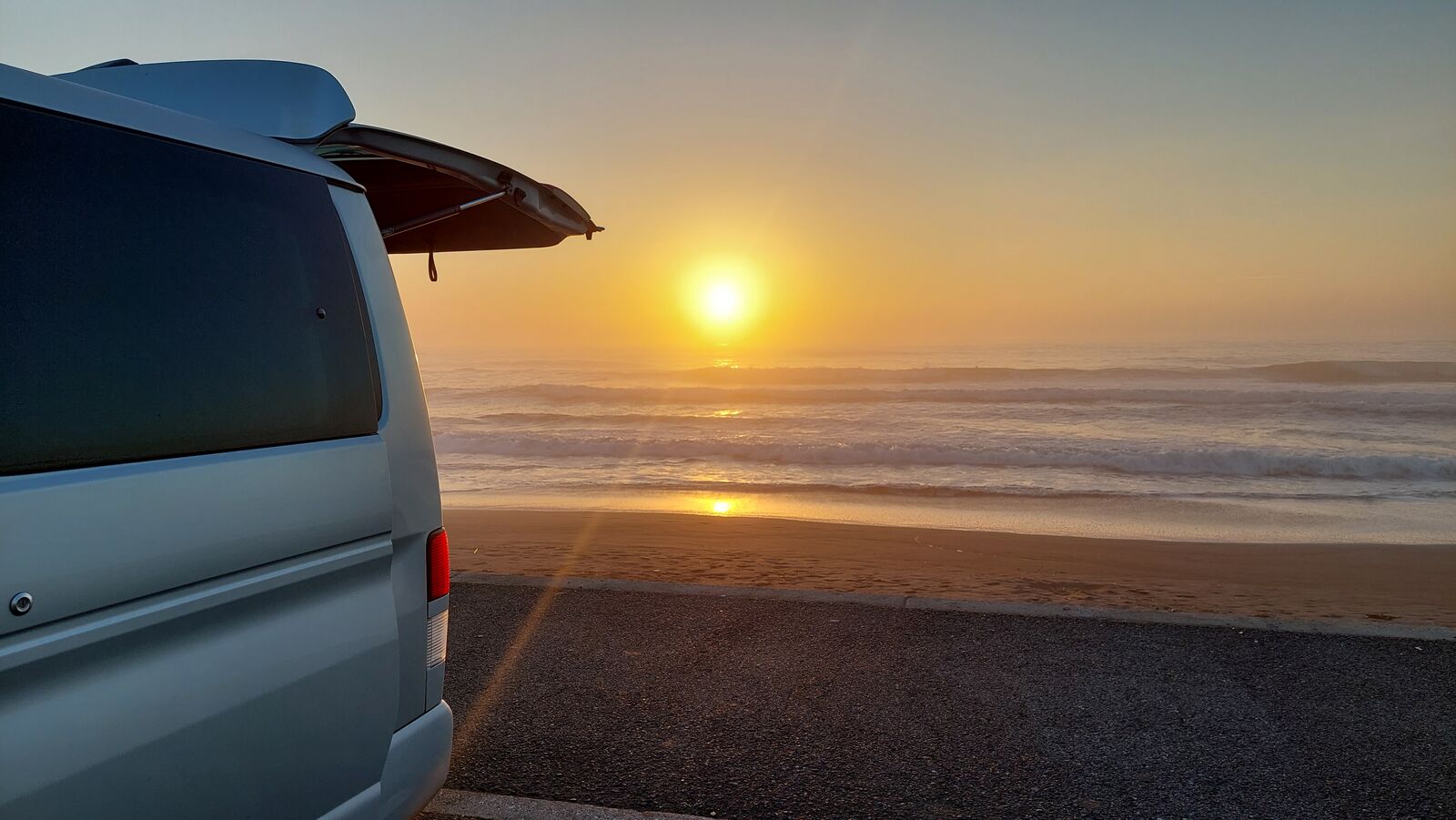 Campervan with open trunk facing Pacific sunrise at Kujukuri Beach, golden light and surfers