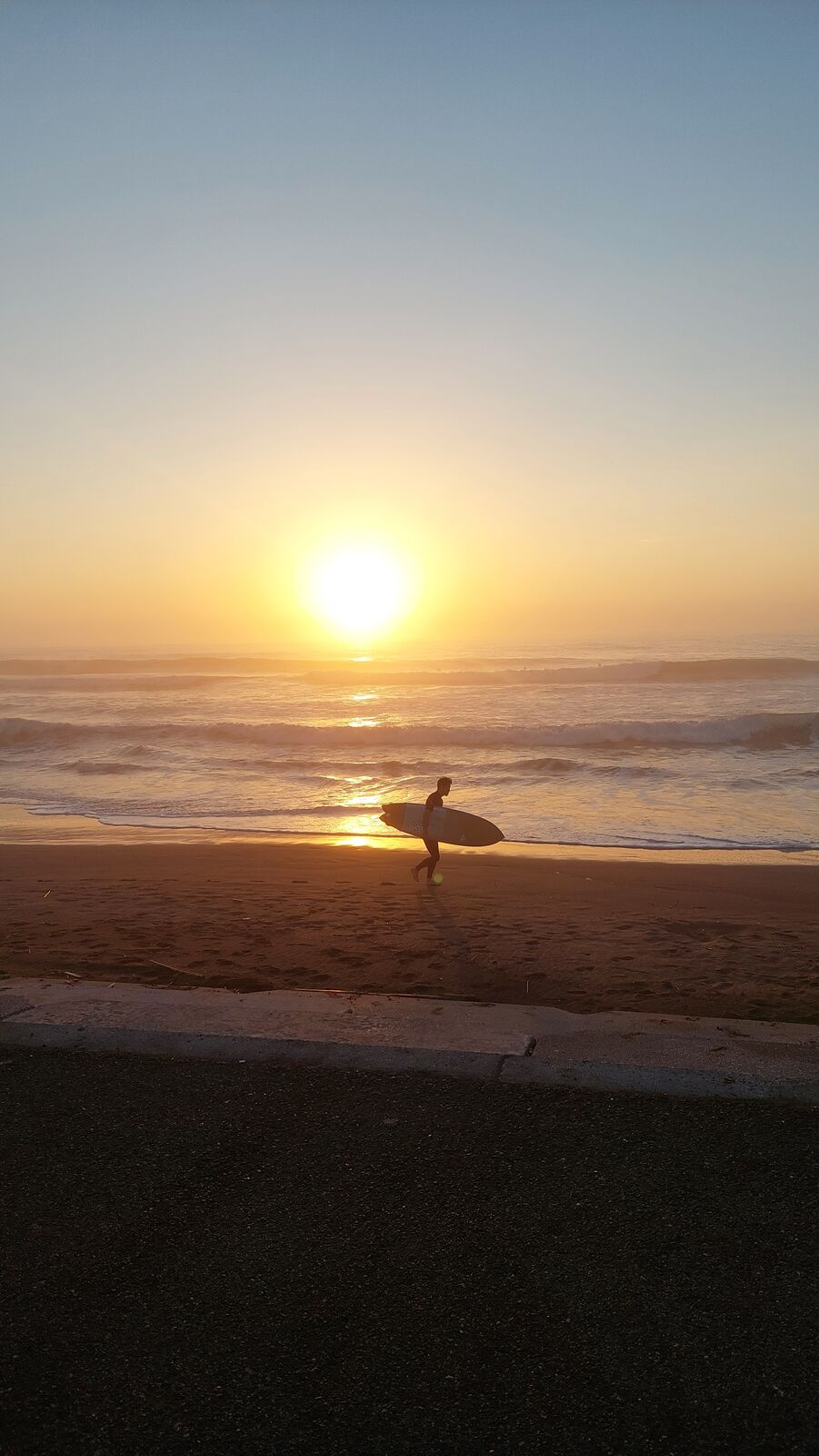 Surfer walking into the sunrise carrying a surfboard, silhouetted against golden light reflecting off the Pacific