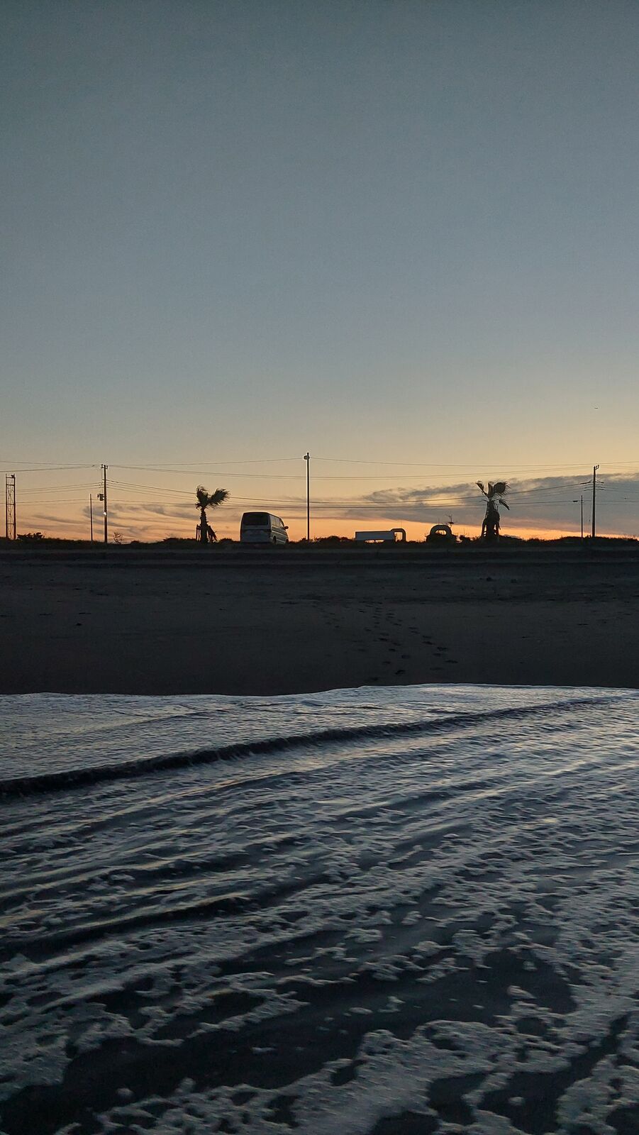 Dusk at the beach: vans and palm trees silhouetted against the last orange glow of sunset, seen from the waterline