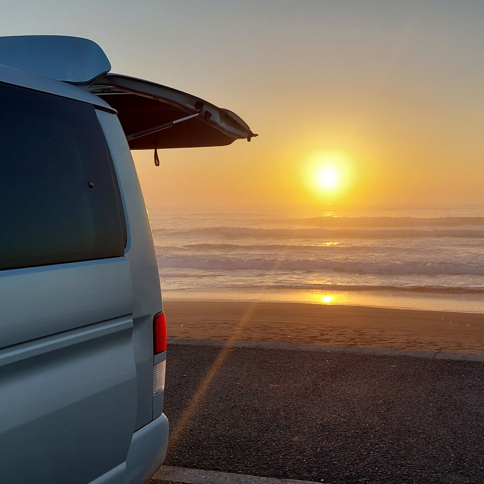 Campervan with open trunk framing the sunrise over the ocean, golden hour light and Pacific waves