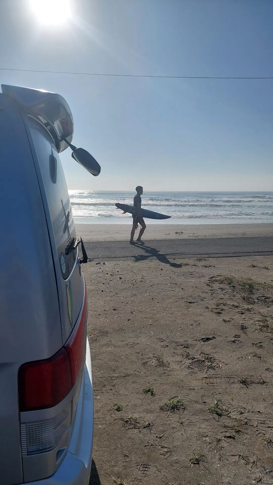 Van parked at the beach with a surfer heading toward the water, clear blue sky morning