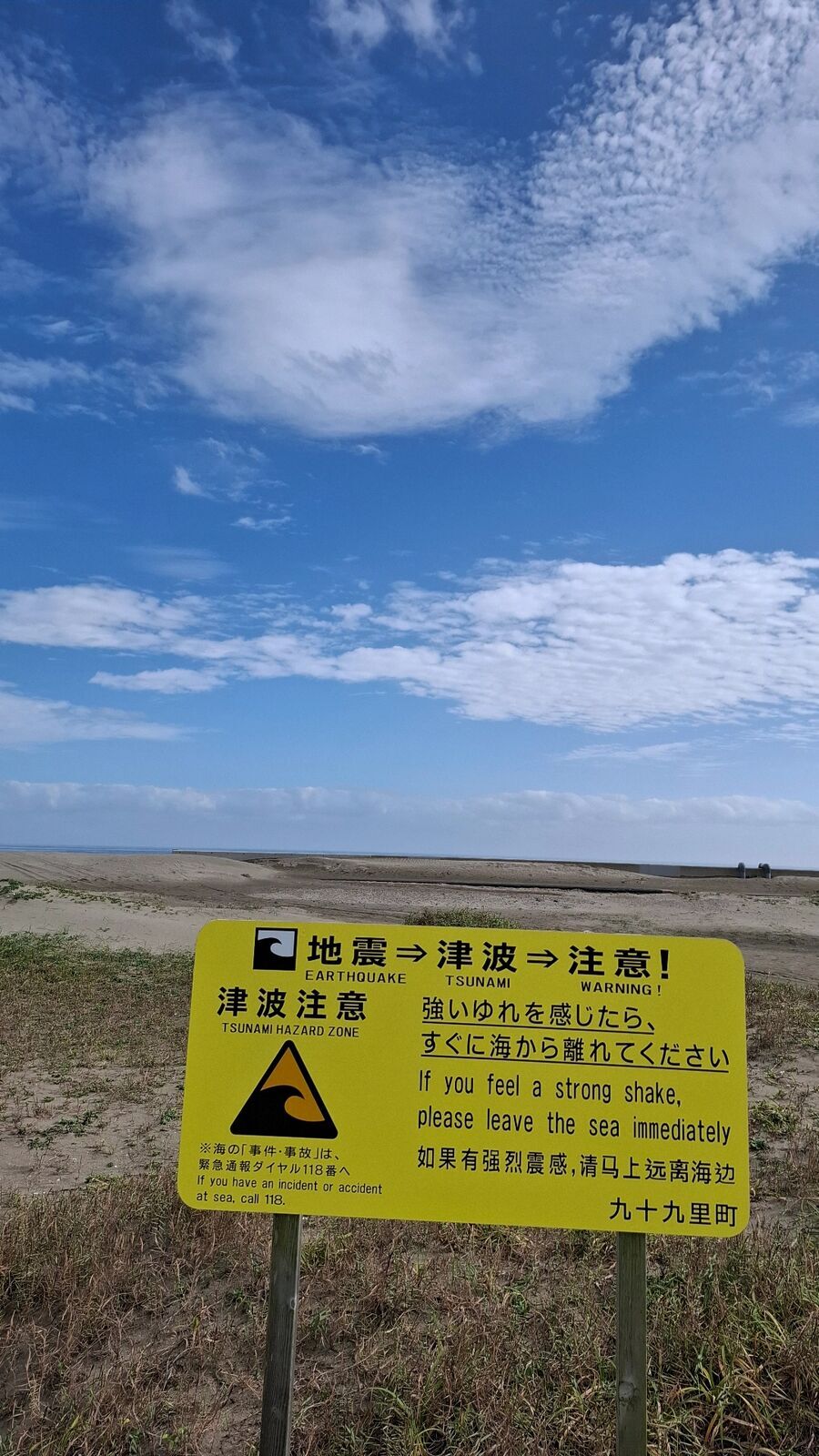Yellow tsunami warning sign reading Kujukuri-machi with the beach and dunes behind under blue sky