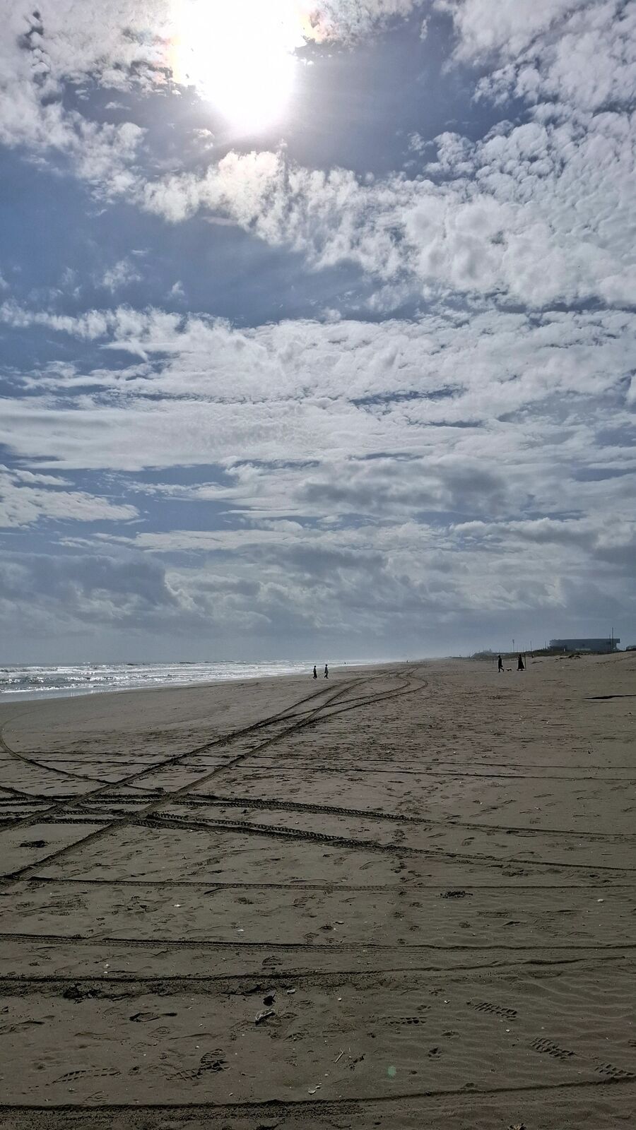 Wide empty beach with tire tracks in the sand, dramatic cloud formations and sun breaking through, two distant figures walking