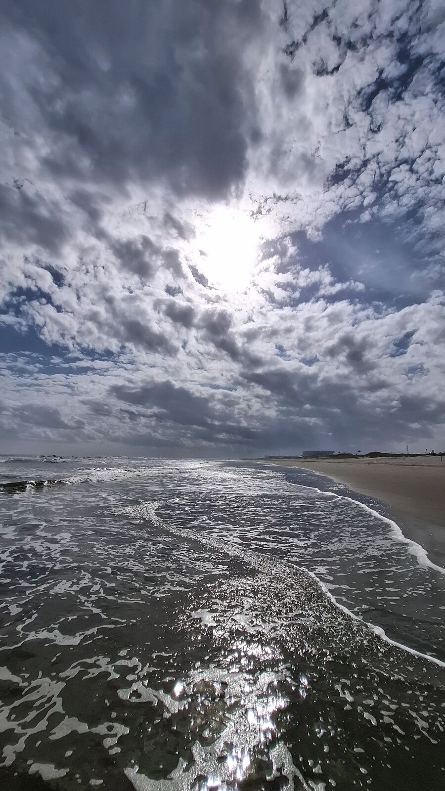 Sun breaking through massive clouds over the Pacific, waves washing up on dark sand, silver light on the water