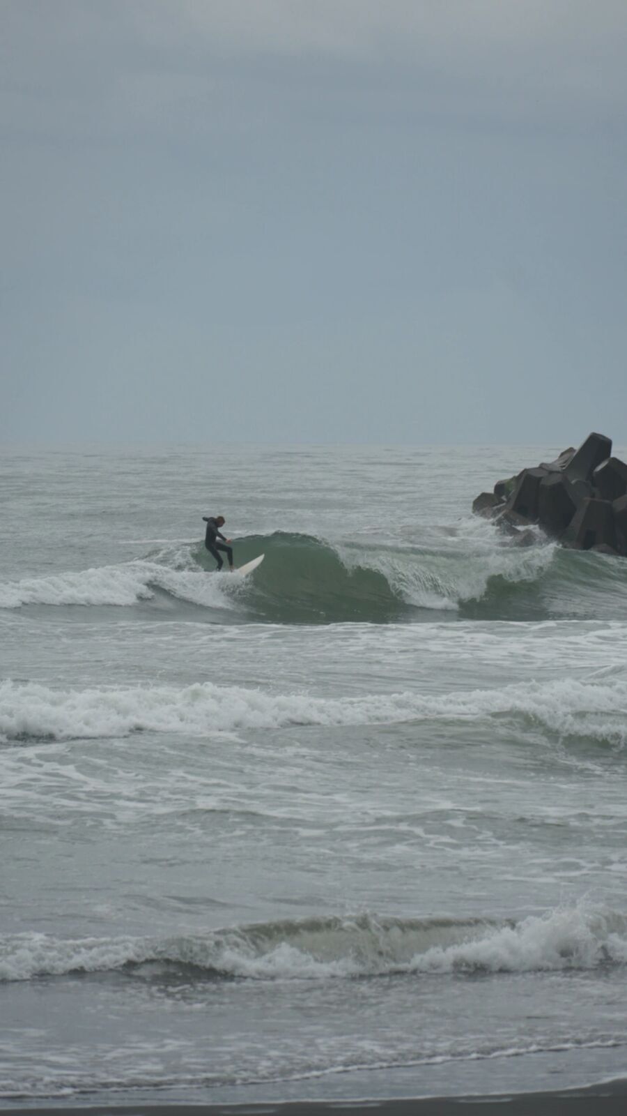 Surfer catching a wave near concrete tetrapod breakwater, grey-green Pacific water with real power behind the swell