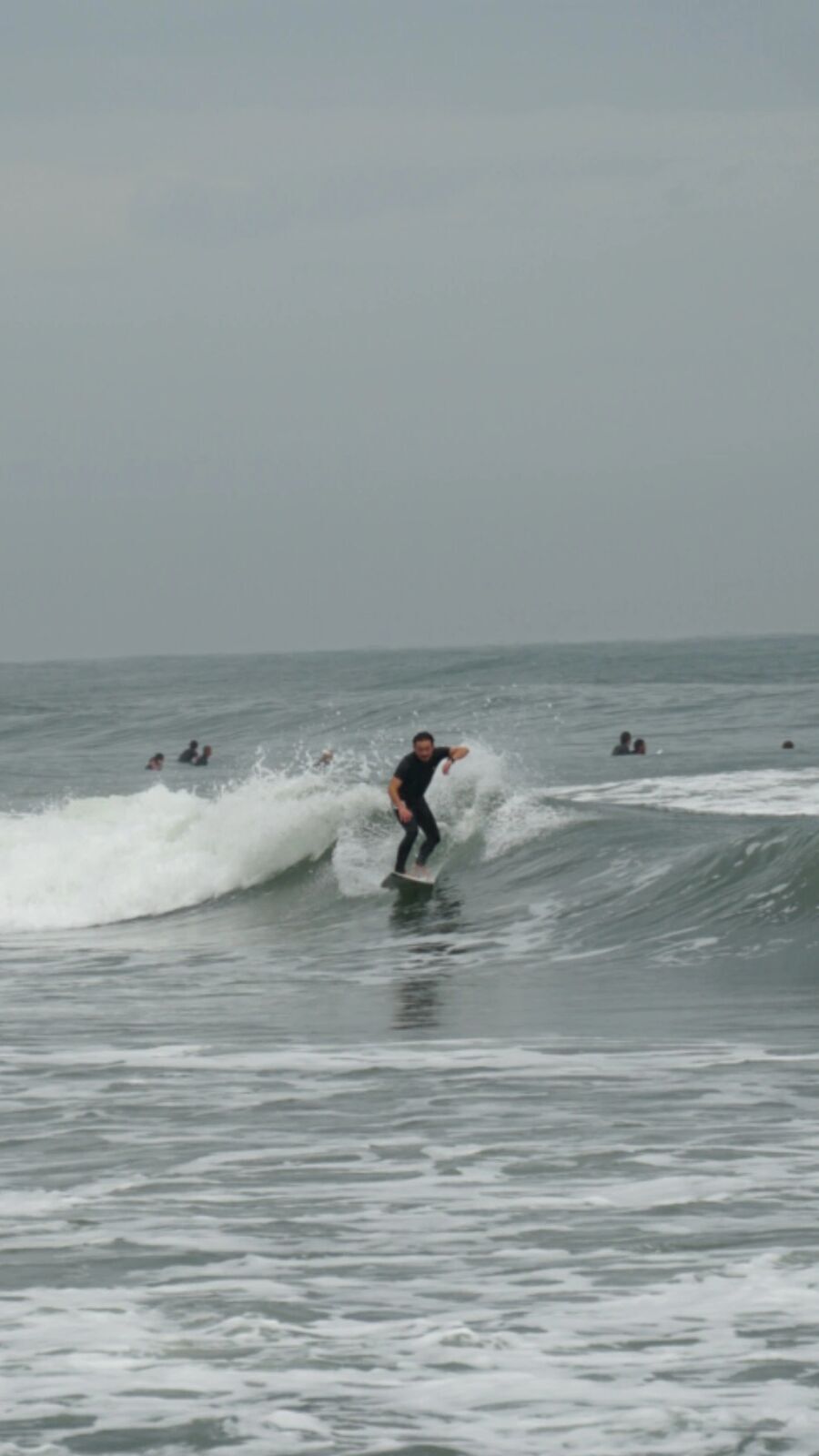 Surfer in black wetsuit riding a wave with other surfers visible in the background, autumn Pacific surf session